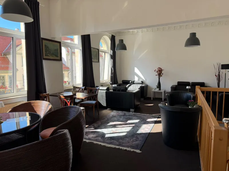 Weiter Blick durch die Lounge: Fensterfront mit Blick auf rote Dächer, Holztische, Ledersofas und ein Teppich im Sonnenlicht